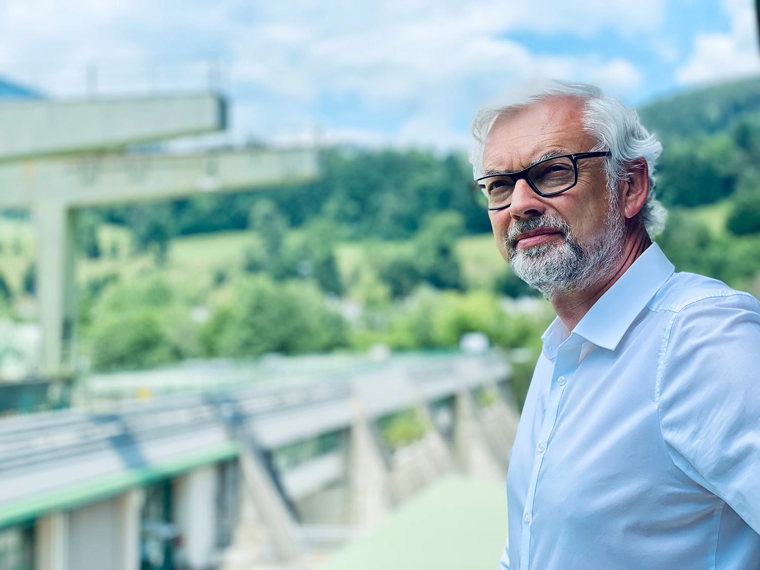 Michael Strugl im hellblauen Hemd. Er blick in die Ferne. Im Hintergrund sehen wir ein Wasserkraftwerk.