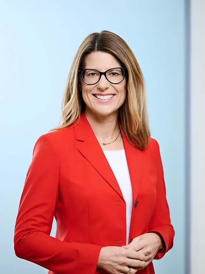 Barbara Schmidt trägt einen roten Blazer mit einer dezenten weißen Bluse darunter. Sie hat langes, mittellanges blondes Haar. Sie trägt eine schwarze Brille. Sie lächelt in die Kamera.