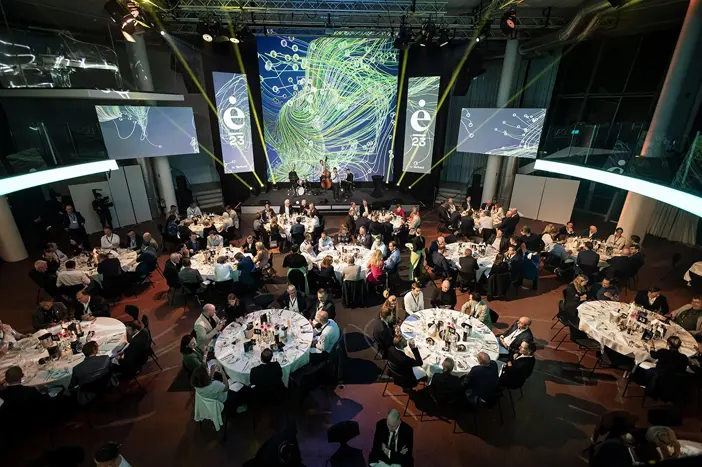 Wir blicken von einer leicht erhöhten Position auf den Dinnerbereich des inspire summits. Auf der Bühne spielt eine Band. Die Teilnehmer sitzen an runden Tischen.