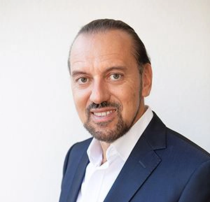 Werner Hoffmann trägt ein weißes Hemd und ein dunkelblaues Sakko. Seine Haar sind streng zurück gekämmt. Die Augen sind braun.