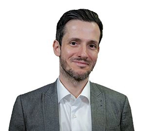 Wolker Schaffler trägt einen Dreitagebart, ein weißes Hemd, ein graues Sakko und ein Lächeln.