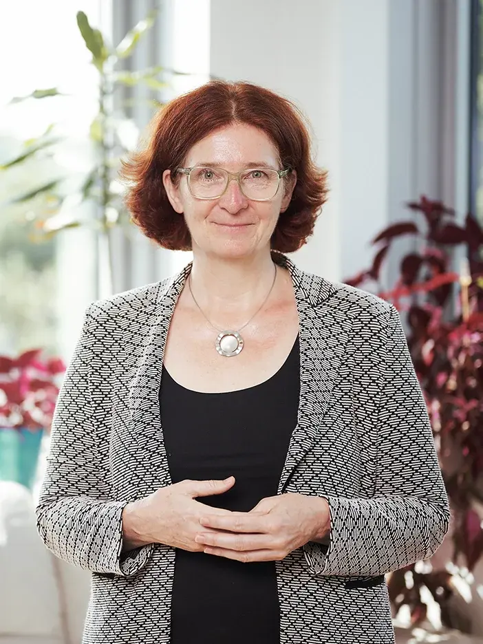 Sigrid Stagl hat rötliches Haar. Sie träg ein schwarzes Oberteil und einen graugemusterten Blazer. Sie lächelt in die Kamera.